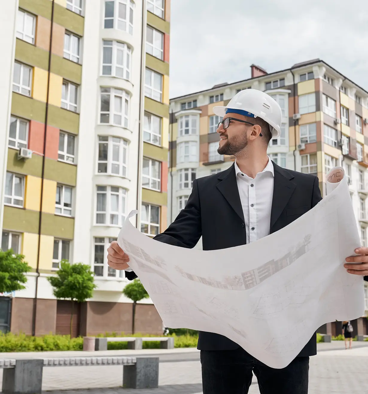 Man in a white hard hat and black suit holding blueprints and looking to the side in front of colorful apartment buildings.